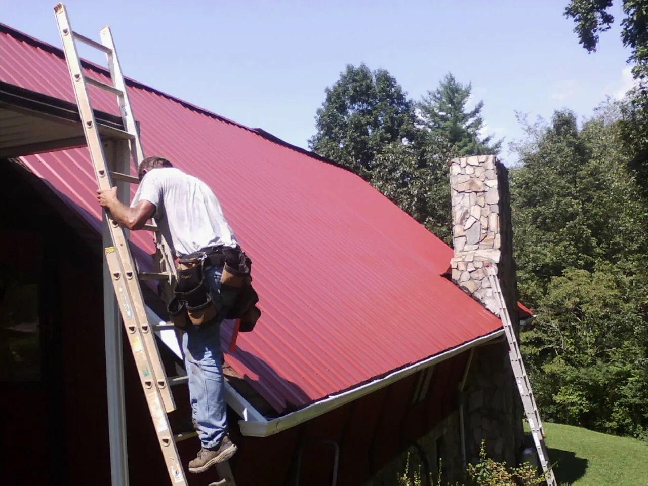 Roofer on a ladder beside a red metal roof and stone chimney — Soffit & Fascia Repair by Roofing Brothers in Antonito